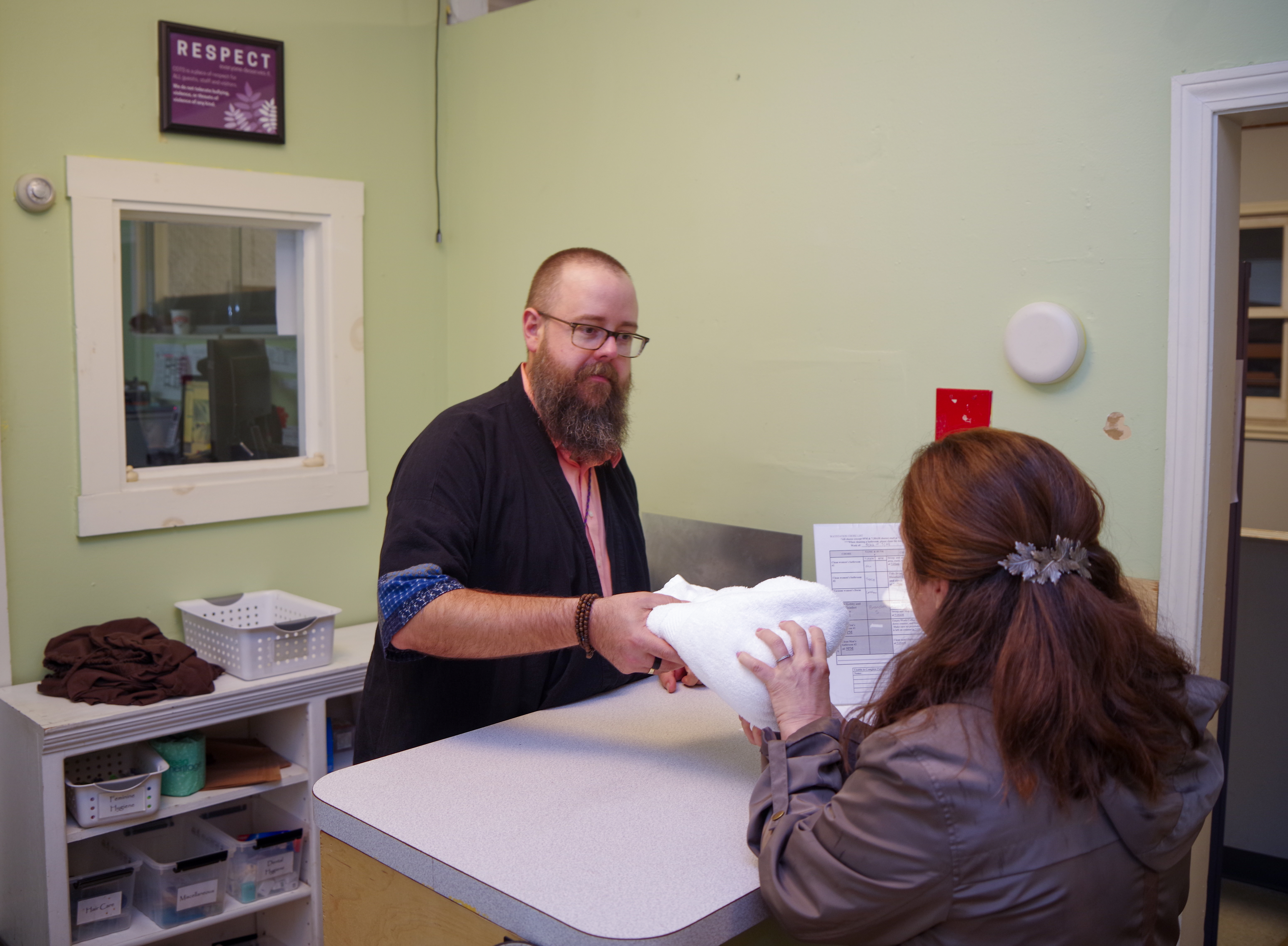 Male COTS staff member with a beard and glasses hands a woman a white towel over a counter.